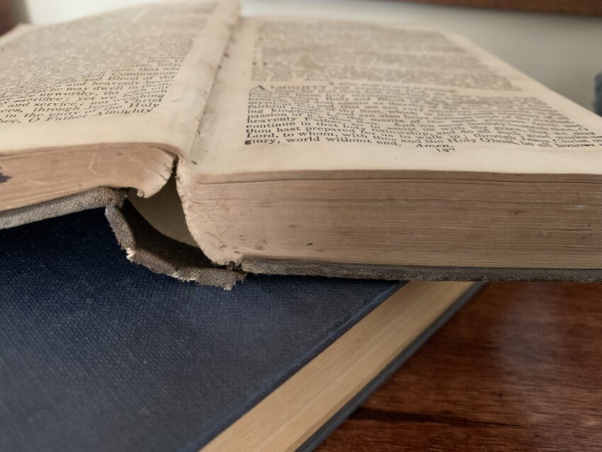 A grey, cloth-bound book rests open atop a blue, cloth-bound book, which in turn rests upon a mahogany wood table. You can tell that it will smell of old books. The pages are aged and the top of the text fades into the background. The bottom two lines are decipherable: "Lord, to whom, with thee and the Holy Ghost, be all honor and glory, world without end. Amen."