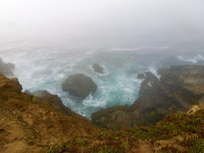 Looking down from the top of a cliff toward the ocean below, where waves splash against a boulder. Fog obscures the horizon and dampens the colors.