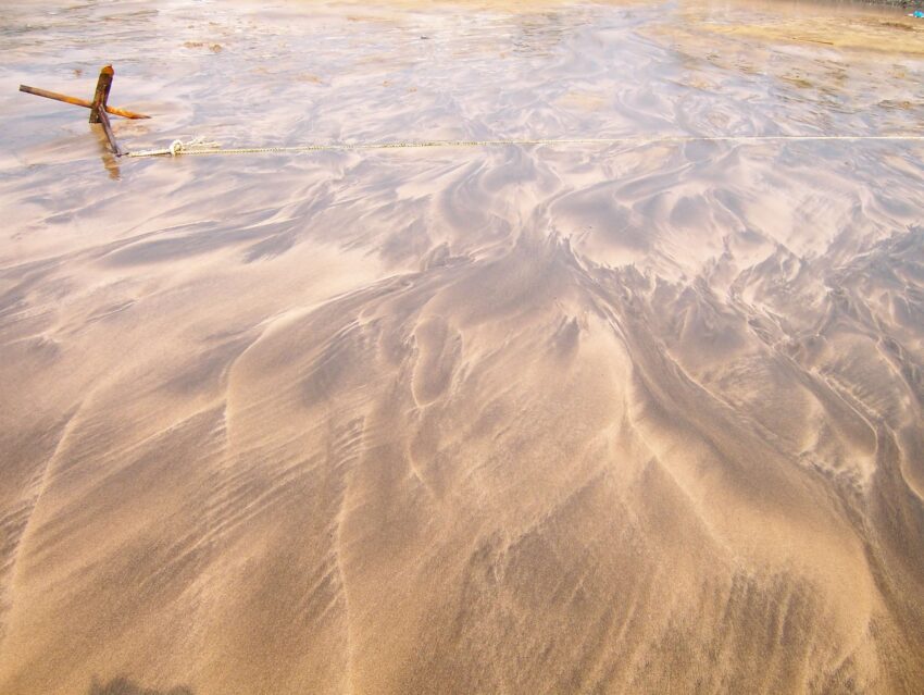 Wavy patterns of wet sand, from dark brown to light tan and shades between, left behind when the tide went out. A rusted anchor rests at the top left of the frame, is rope taut across the top of the picture.